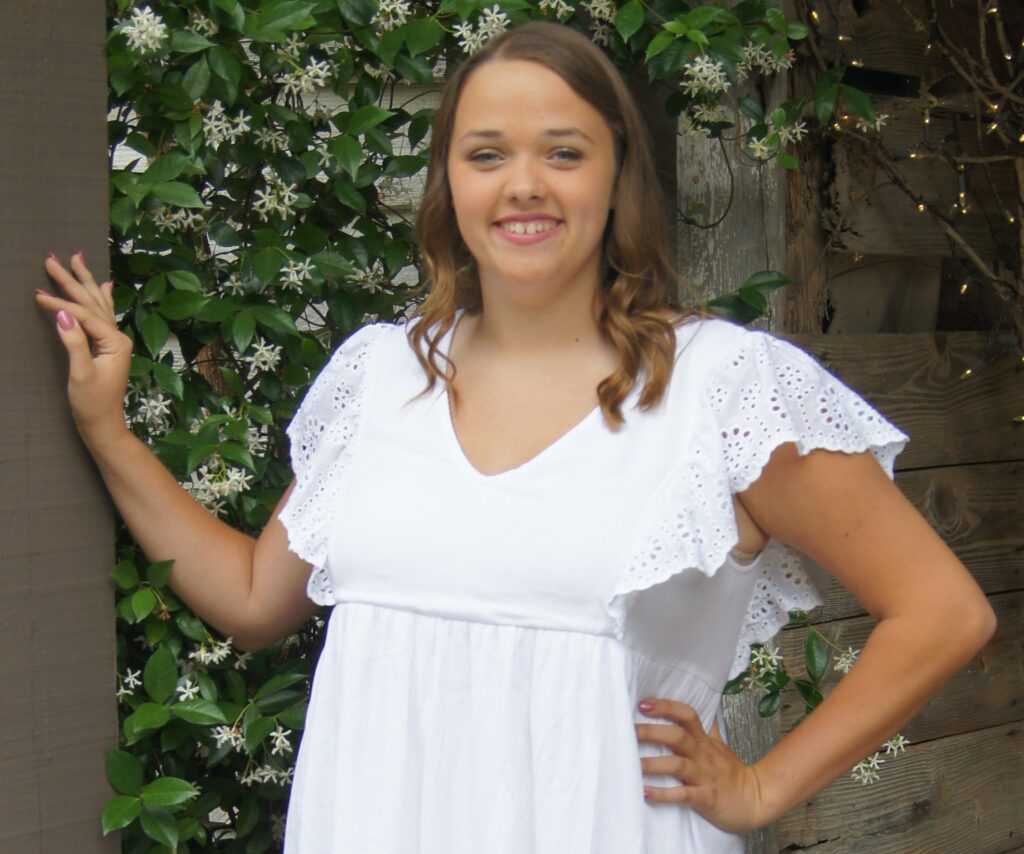 A woman in a white lace dress standing in front of a blooming plant and barn.