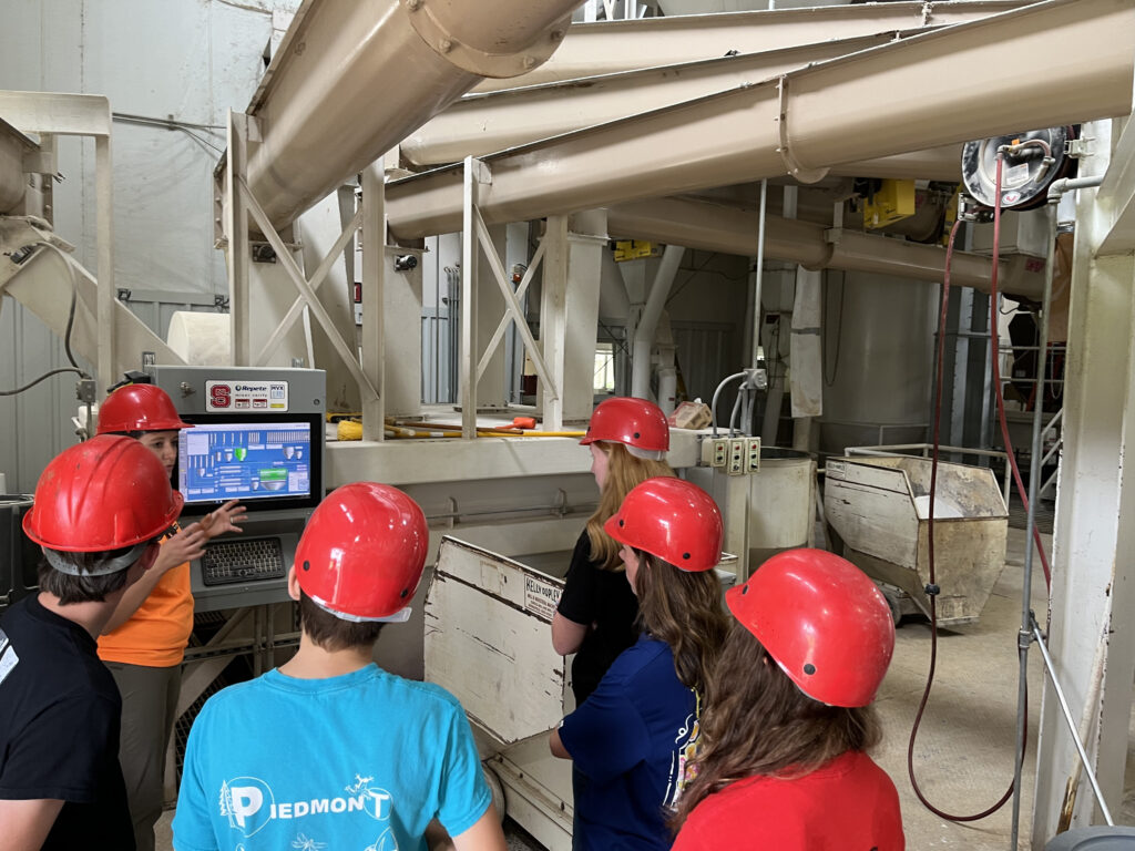 Teenagers wearing red hard hats tour the inside of a feedmill.