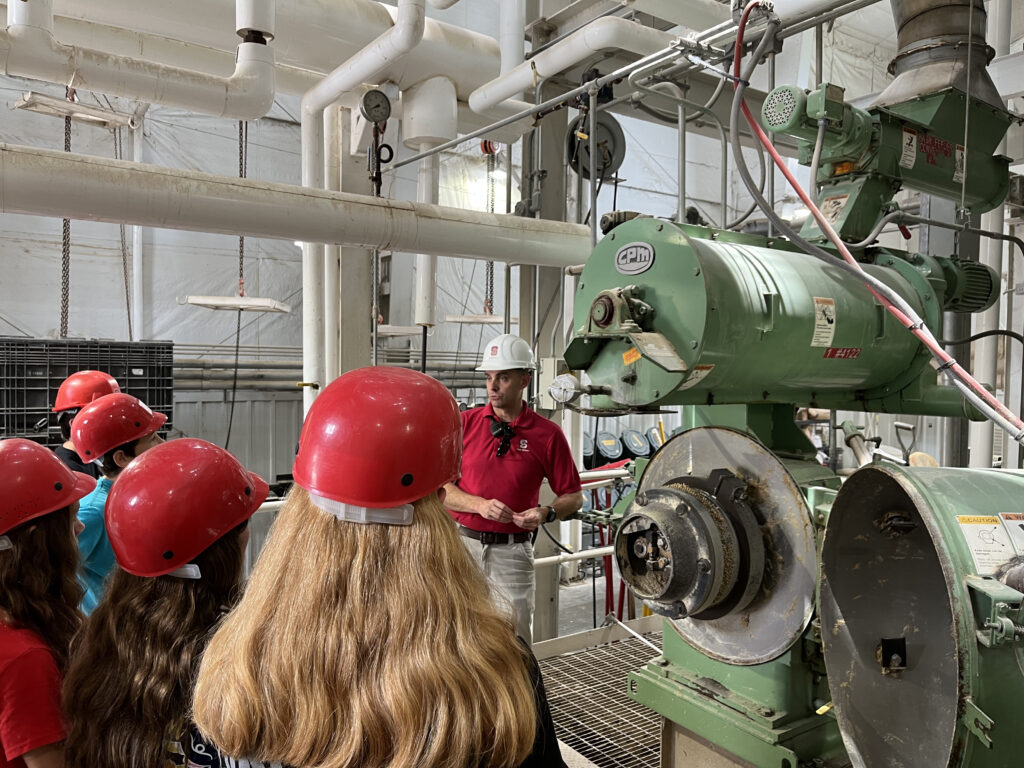 An employee in a hardhat discuss heavy machinery inside a feedmill with students.