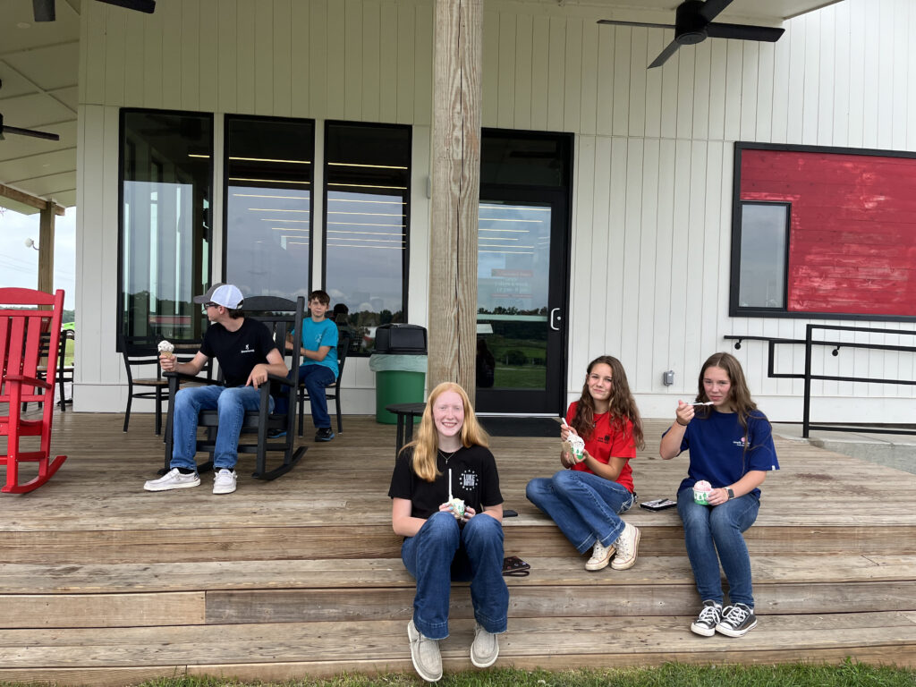A group of teenagers sit on a wood porch eating icecream.