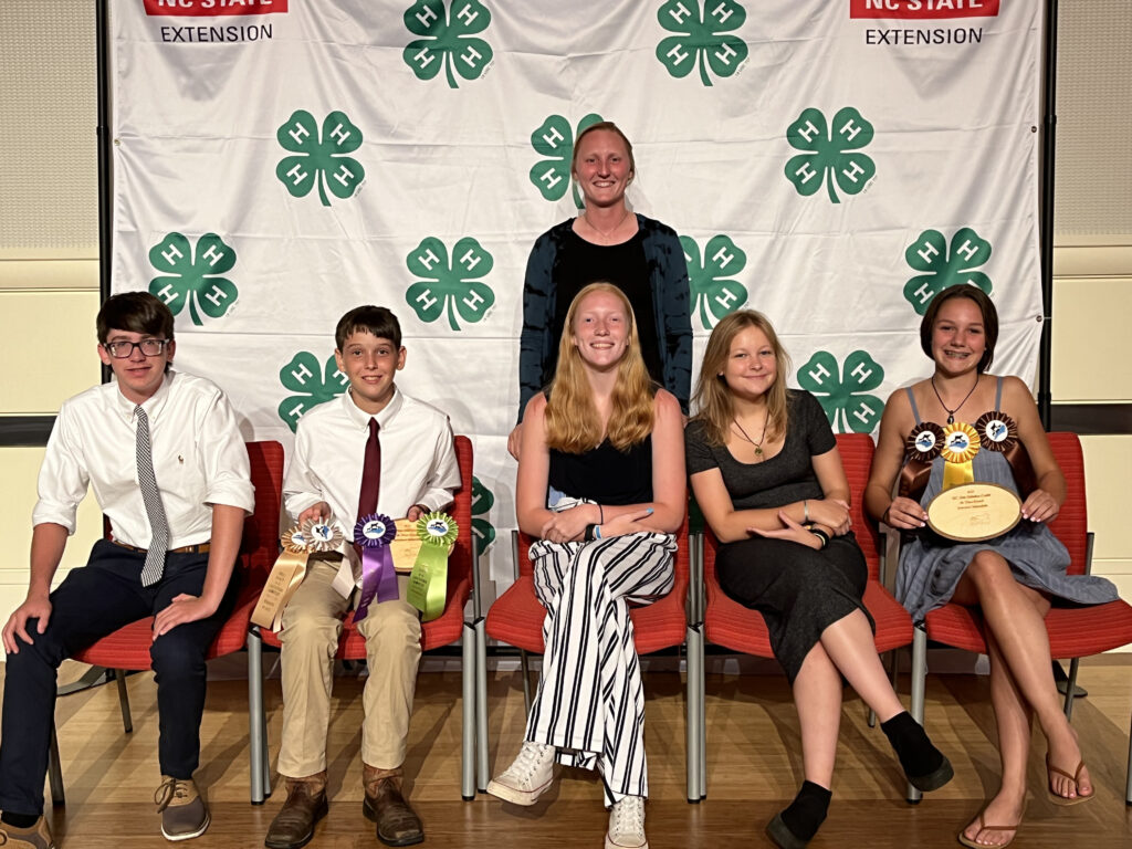 A group of youths posing in front of a 4-H Banner. The group is displaying a number of ribbons and plaques. 