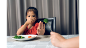 Child covering mouth and holding a hand out to refuse broccoli being held by an adult.